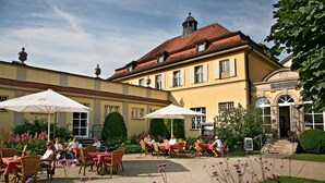 An outdoor café with tables and chairs, surrounded by a garden with flowers and a building with a red roof in the background. - Badhotel Bad Brückenau (Bad Brueckenau)