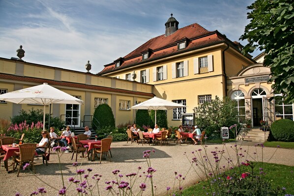 An outdoor café with tables and chairs, surrounded by a garden with flowers and a building with a red roof in the background.