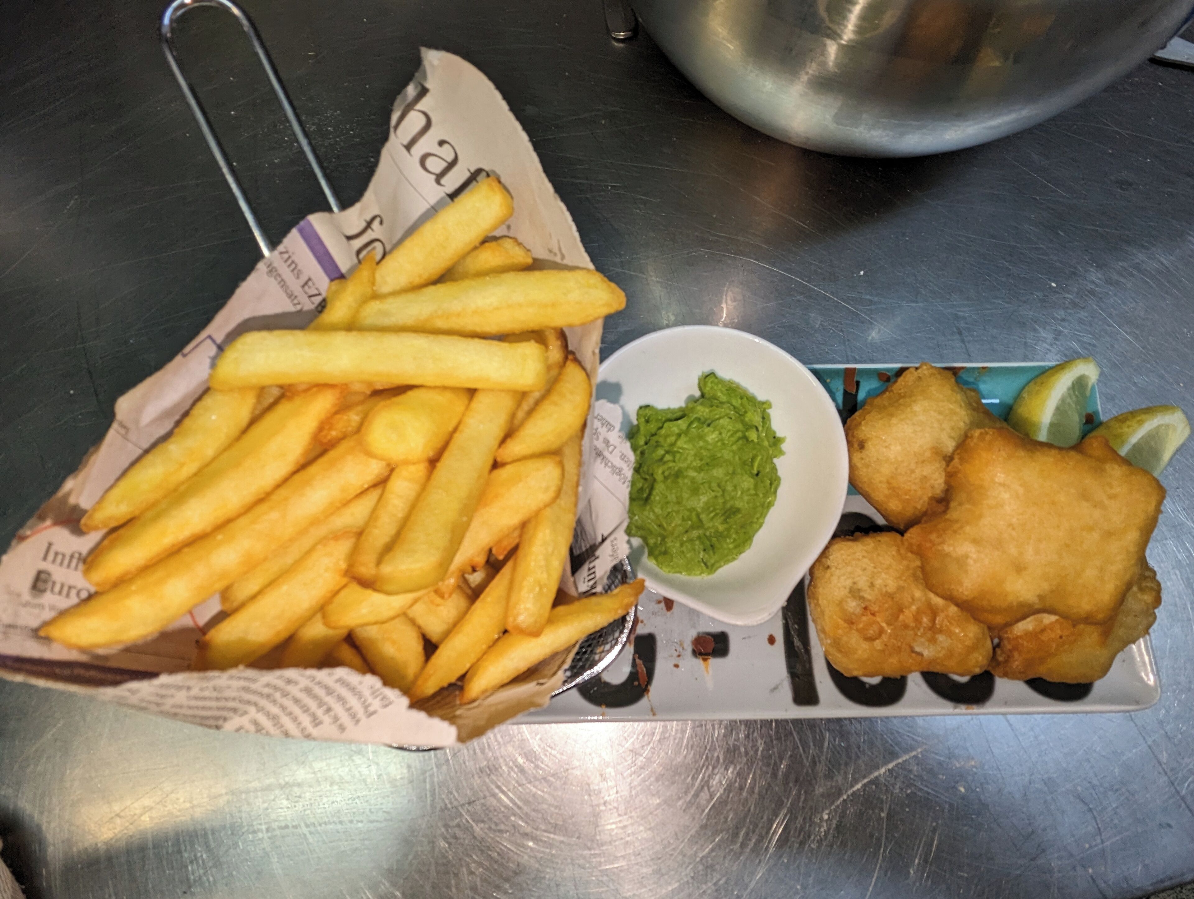 Un repas composé de frites, d’une portion de sauce verte et de poisson pané, servi sur une table en métal.