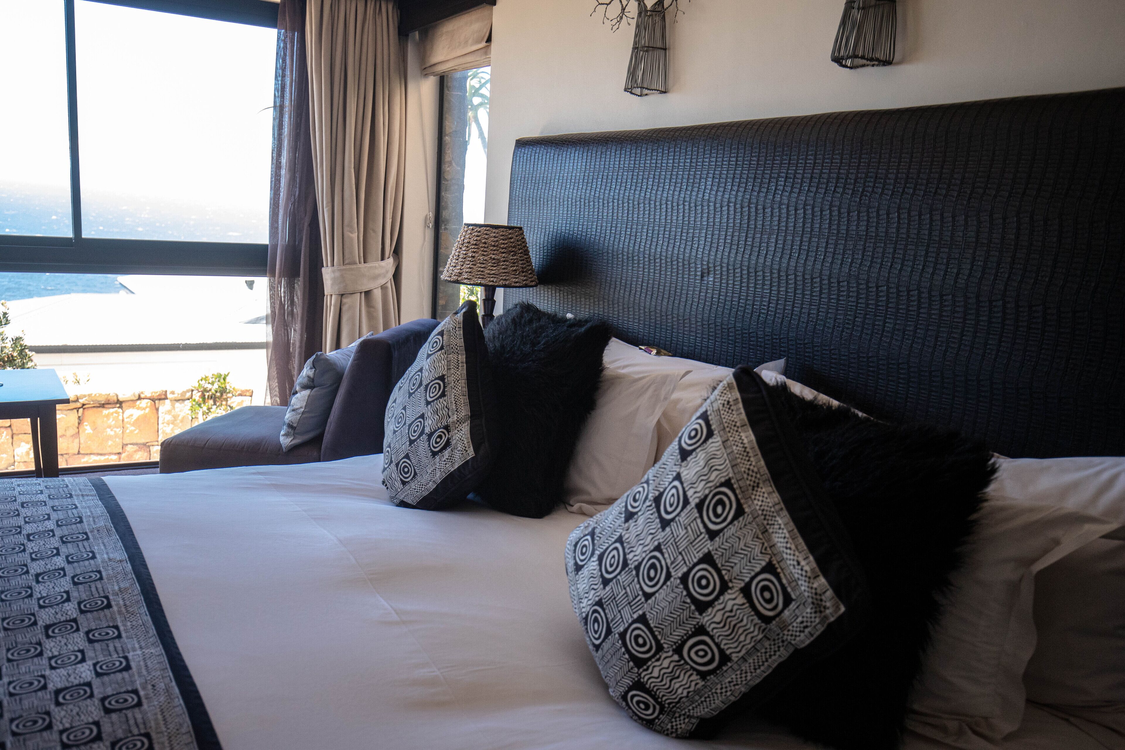A hotel room with a large bed, patterned pillows, a nightstand, and a view of the city.