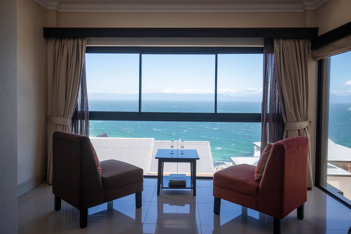 A room with two armchairs and a small table, large windows with curtains, and a view of the sea.
