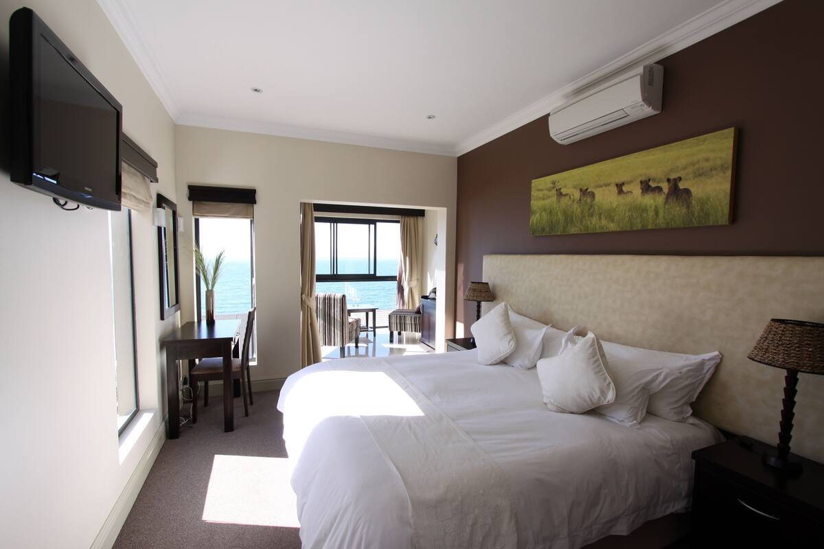 A hotel room with a large bed, a television, a dining area with a table and chairs, and a view of the sea through an open door.