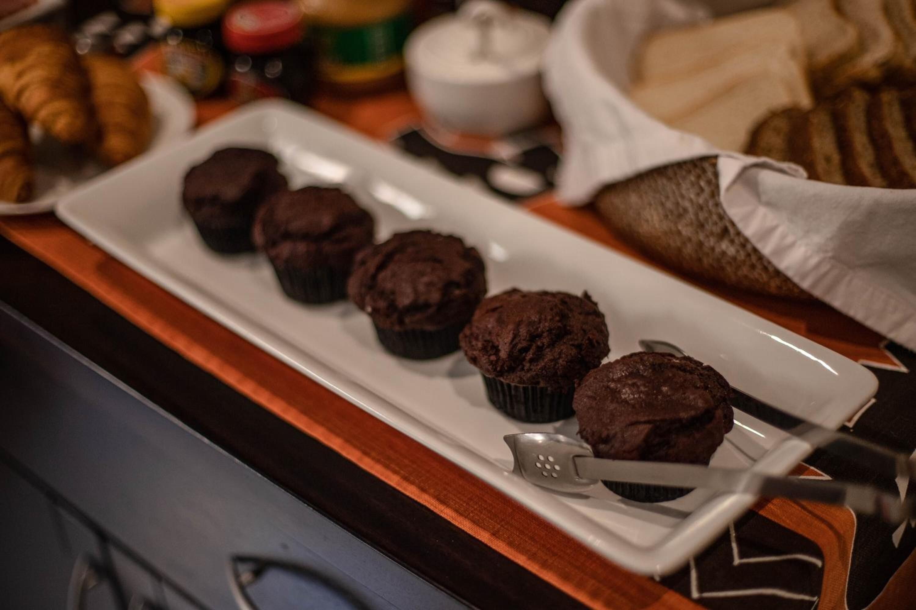 Een schaal met chocolademuffins op een wit bord, met een lepel en een mandje croissants op de achtergrond.