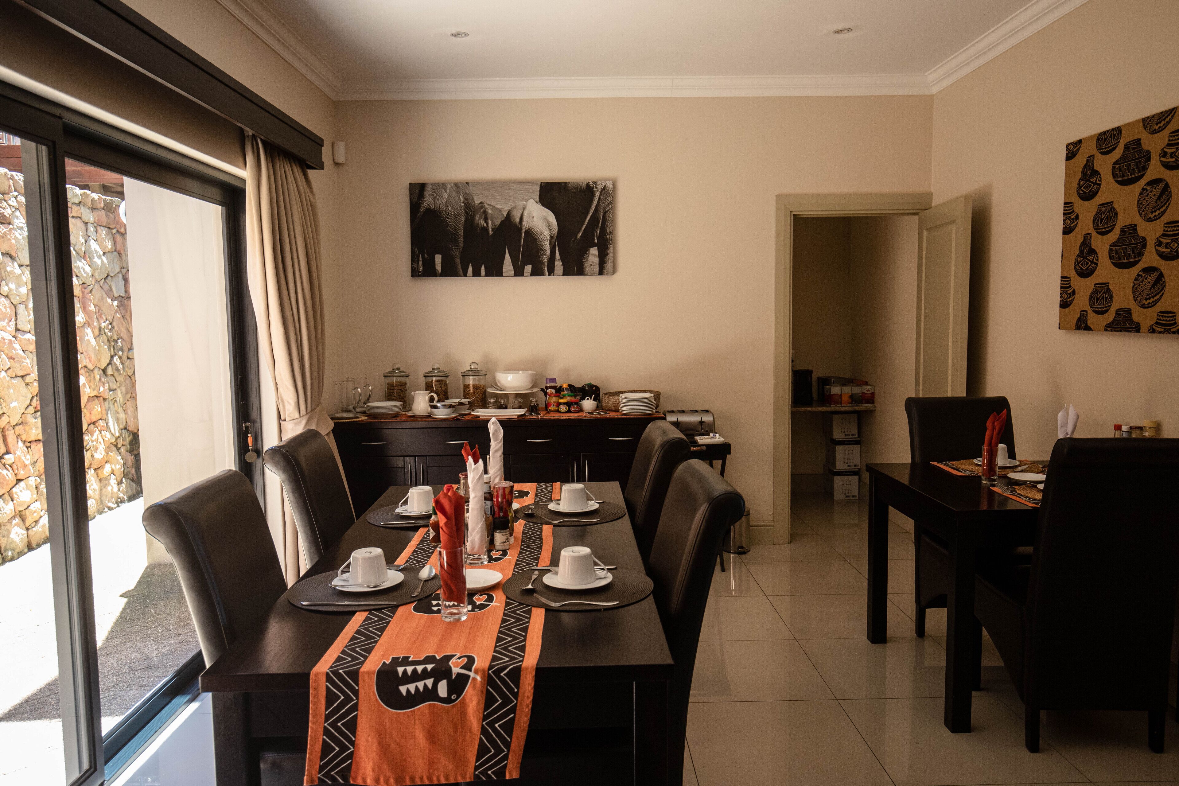 A dining area with a long table set for four, surrounded by black chairs. A large window with curtains is on the left, and a kitchen area is visible in the background.