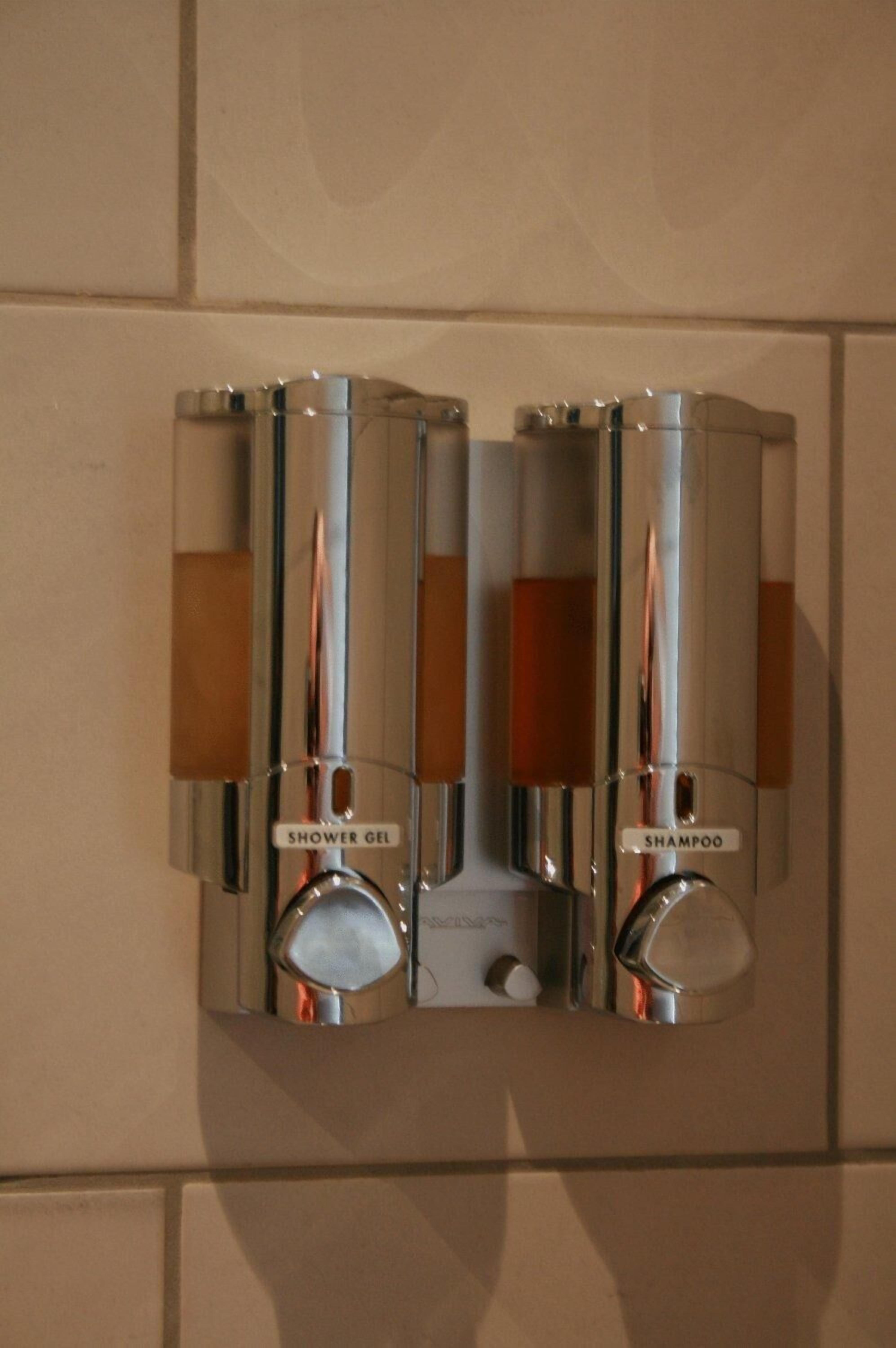 Un distributeur mural à deux compartiments, l’un portant la mention « gel douche » et l’autre « shampoing ».
