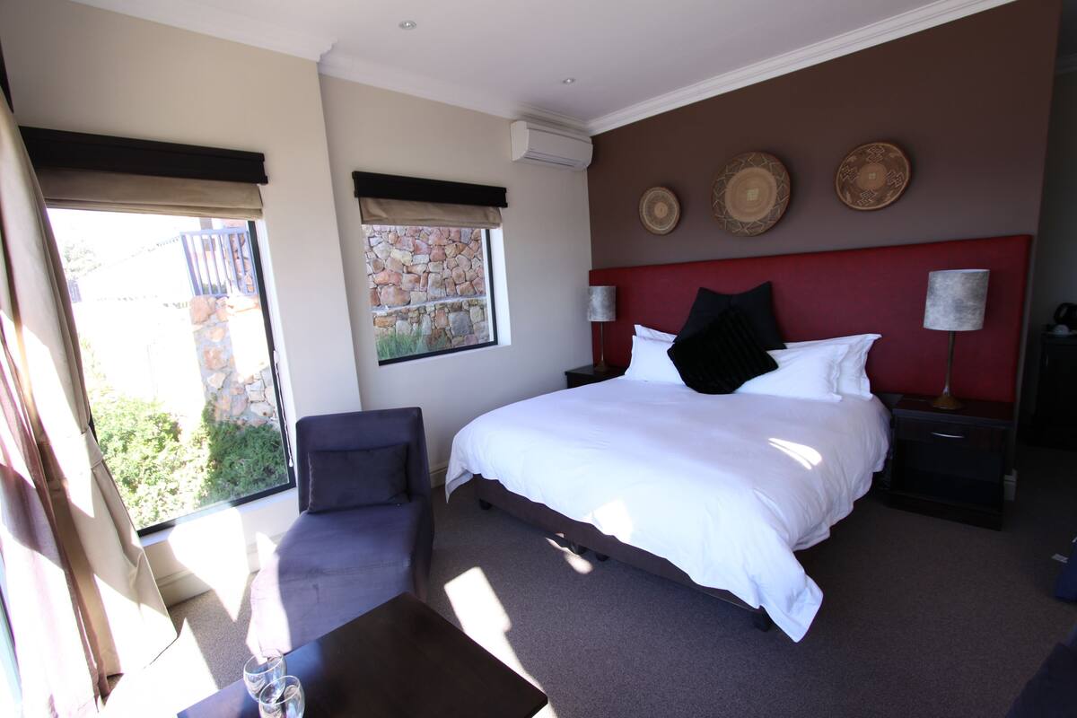A bedroom with a bed, a chair, a nightstand, a lamp, and a window with a view of greenery.