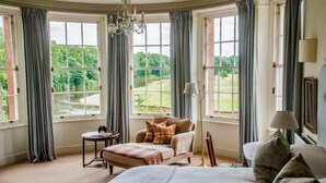 A bedroom with a large bed, a sofa, and a chair. There are multiple windows with curtains, a chandelier, and a lamp. - Warwick Hall (Carlisle)