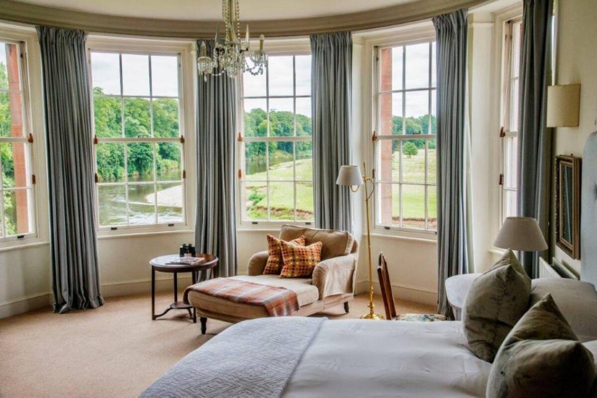 Quarto com uma cama grande, um sofá e uma poltrona. Há várias janelas com cortinas, um candelabro e um abajur.