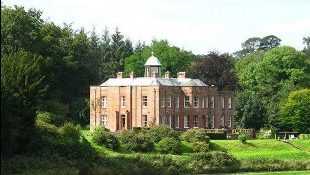 A large brick building with a central tower, surrounded by lush greenery and a river. - Warwick Hall (Carlisle)