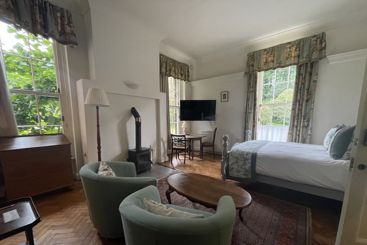 Um quarto com uma cama, duas poltronas, uma mesa de madeira, uma televisão e uma lareira.