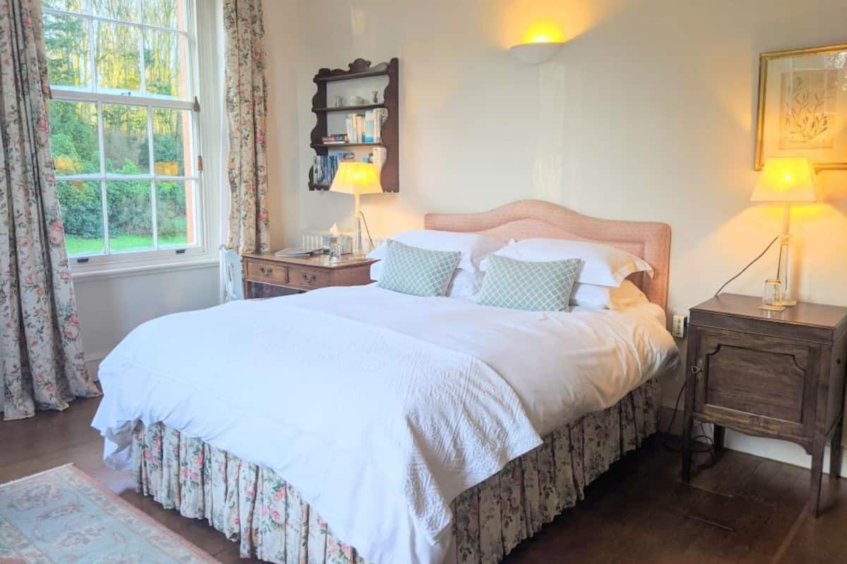 Cama bem arrumada com saia de flores, mesa de cabeceira com abajur e criado-mudo com abajur. Há uma janela com cortinas, prateleira na parede com livros, e um quadro emoldurado.