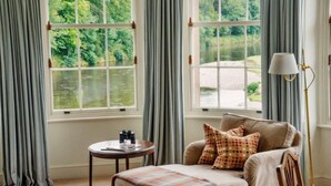 A room with a large window overlooking a river, a sofa with patterned cushions, a small round table with a lamp, and a view of greenery outside. - Warwick Hall (Carlisle)