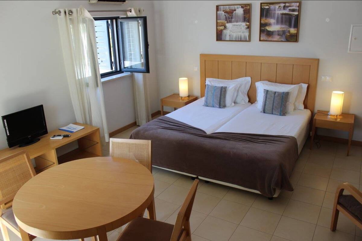 Une chambre d’hôtel avec un lit double, deux tables de chevet avec des lampes, une télévision, une table ronde en bois avec des chaises et une fenêtre avec des rideaux.