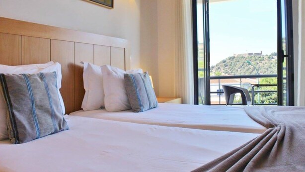 A hotel room with two beds, a wooden headboard, a balcony with a view of a hill and buildings, and a table outside.