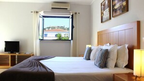 A hotel room with a bed, a television on a wooden stand, a window with curtains, and a wall-mounted air conditioner.