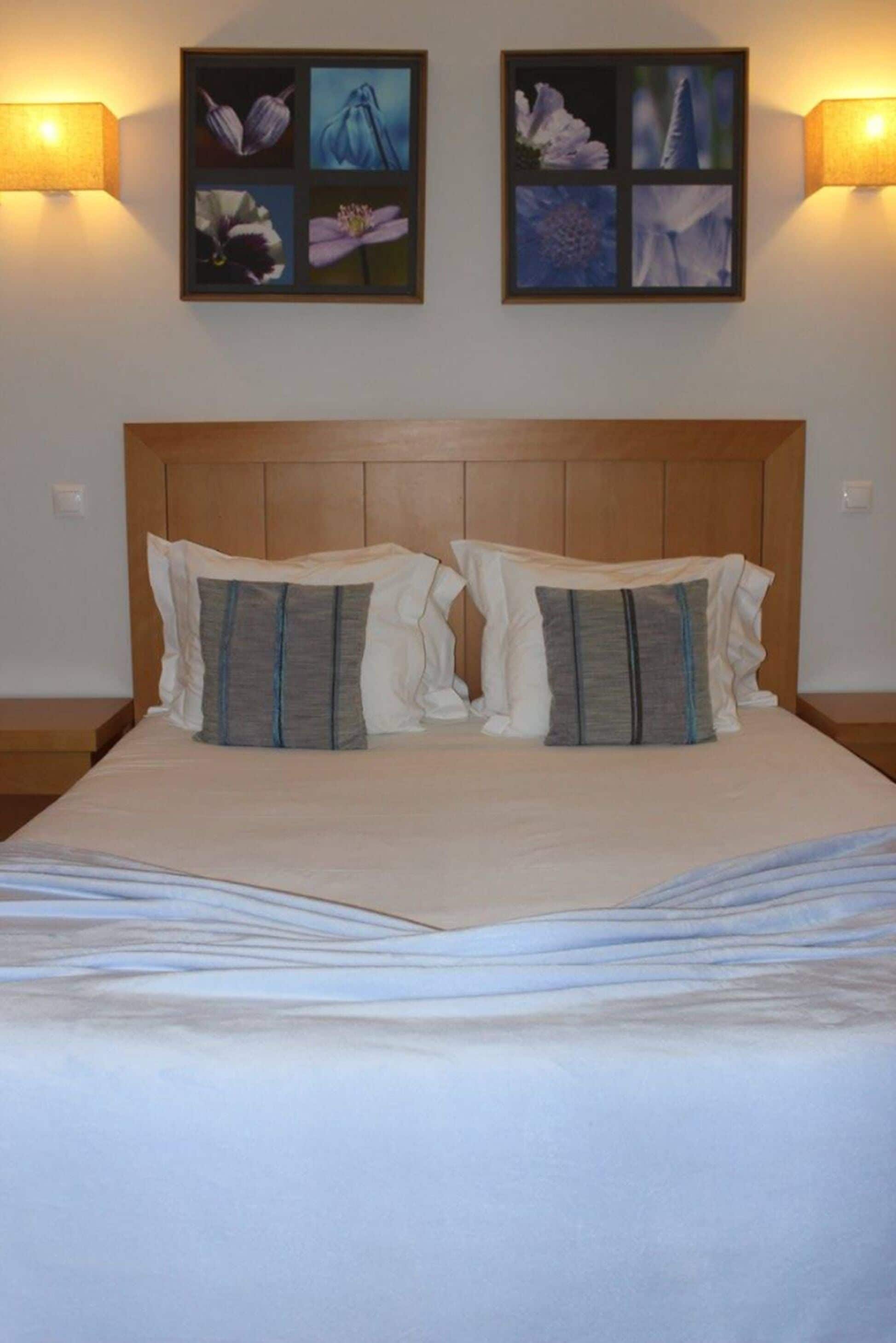 A neatly made bed with a wooden headboard, two wall-mounted lamps, and a floral artwork above the bed.