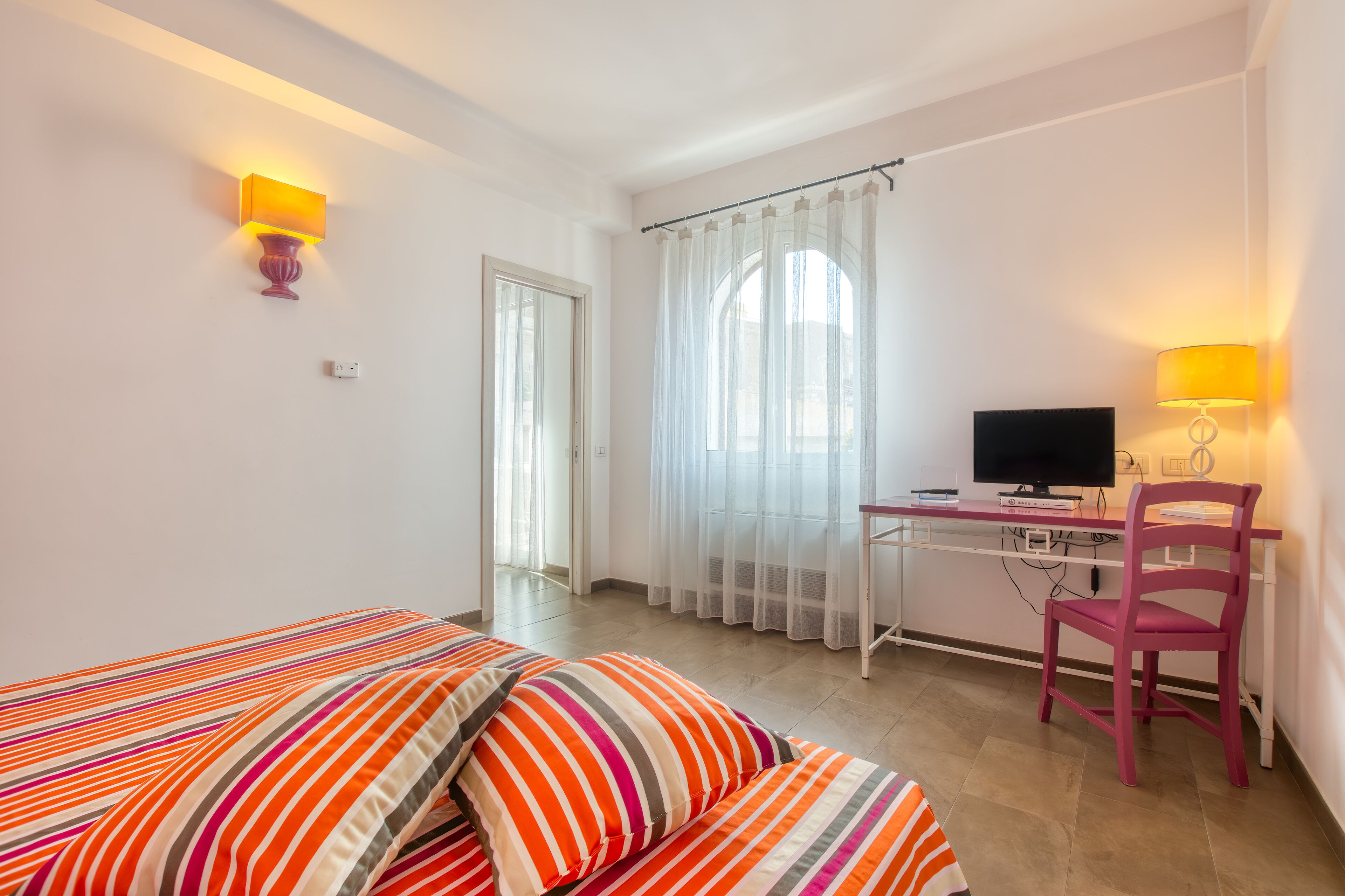 A bedroom with a bed, a desk, a chair, a TV, and a window with curtains.