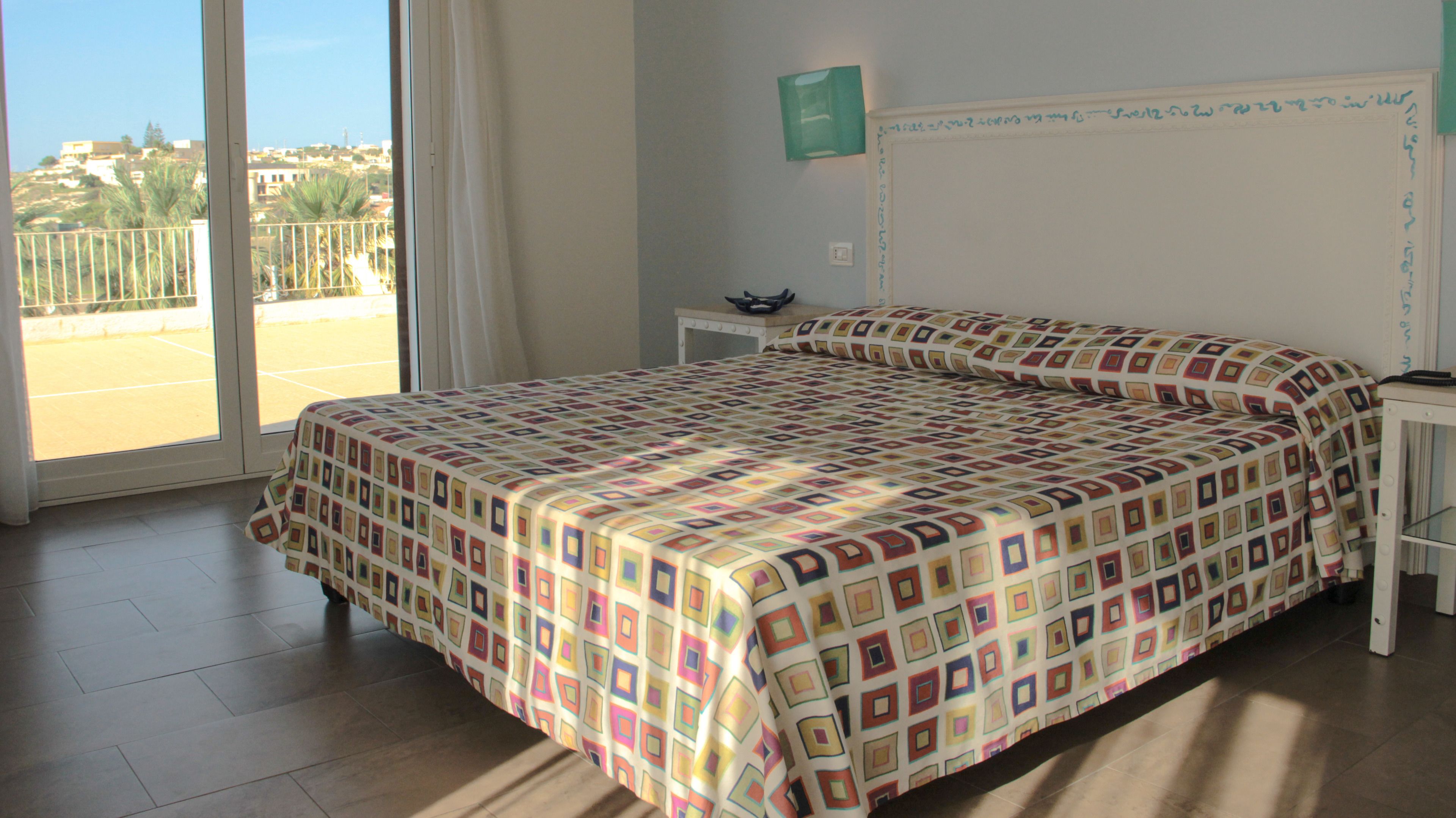 A bedroom with a bed, a nightstand, a window with a view of a tennis court and buildings, and a wall-mounted light fixture.