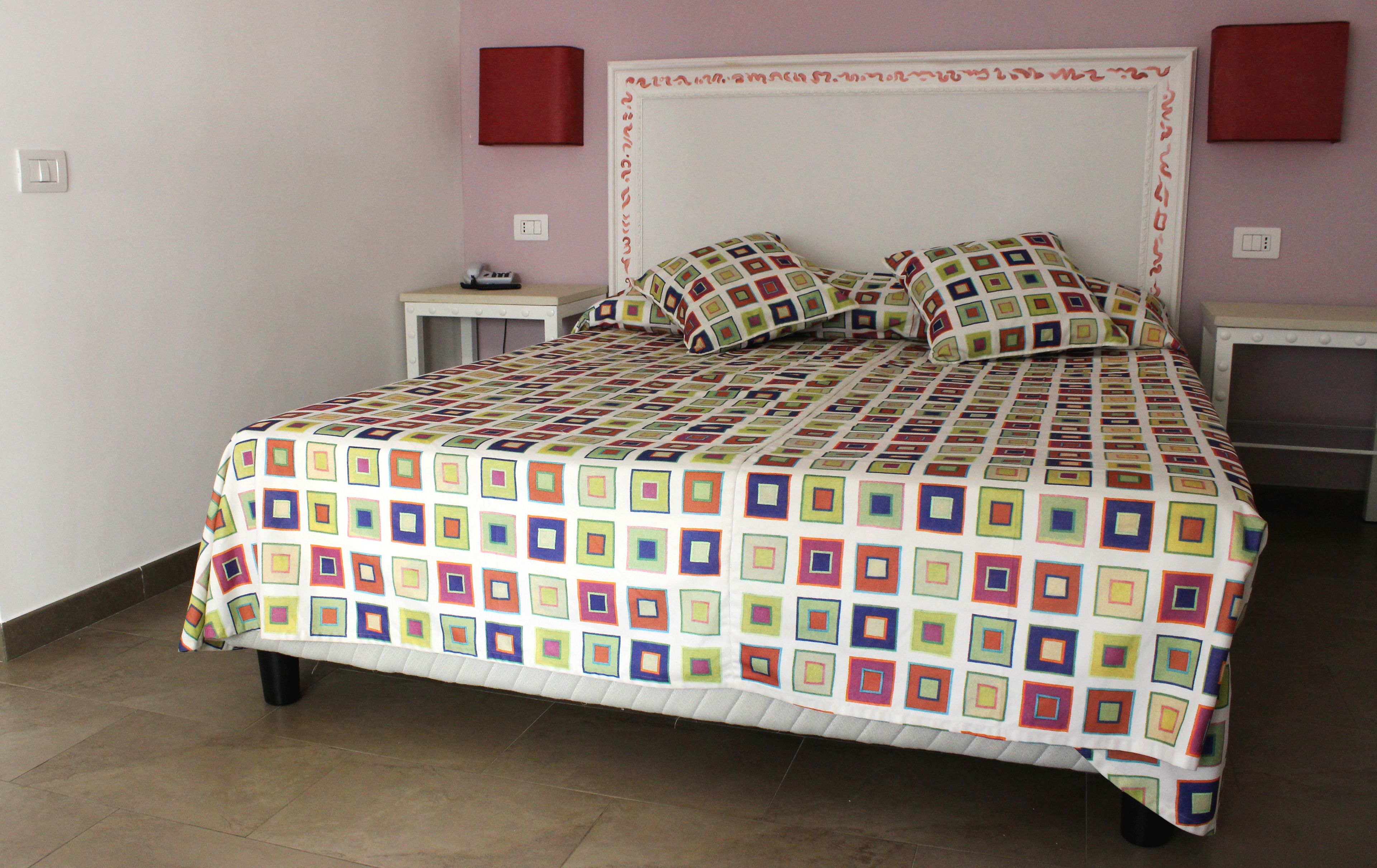 Una cama con un edredón de diseño geométrico y colores vivos, un cabecero blanco y dos mesitas de noche con lámparas.