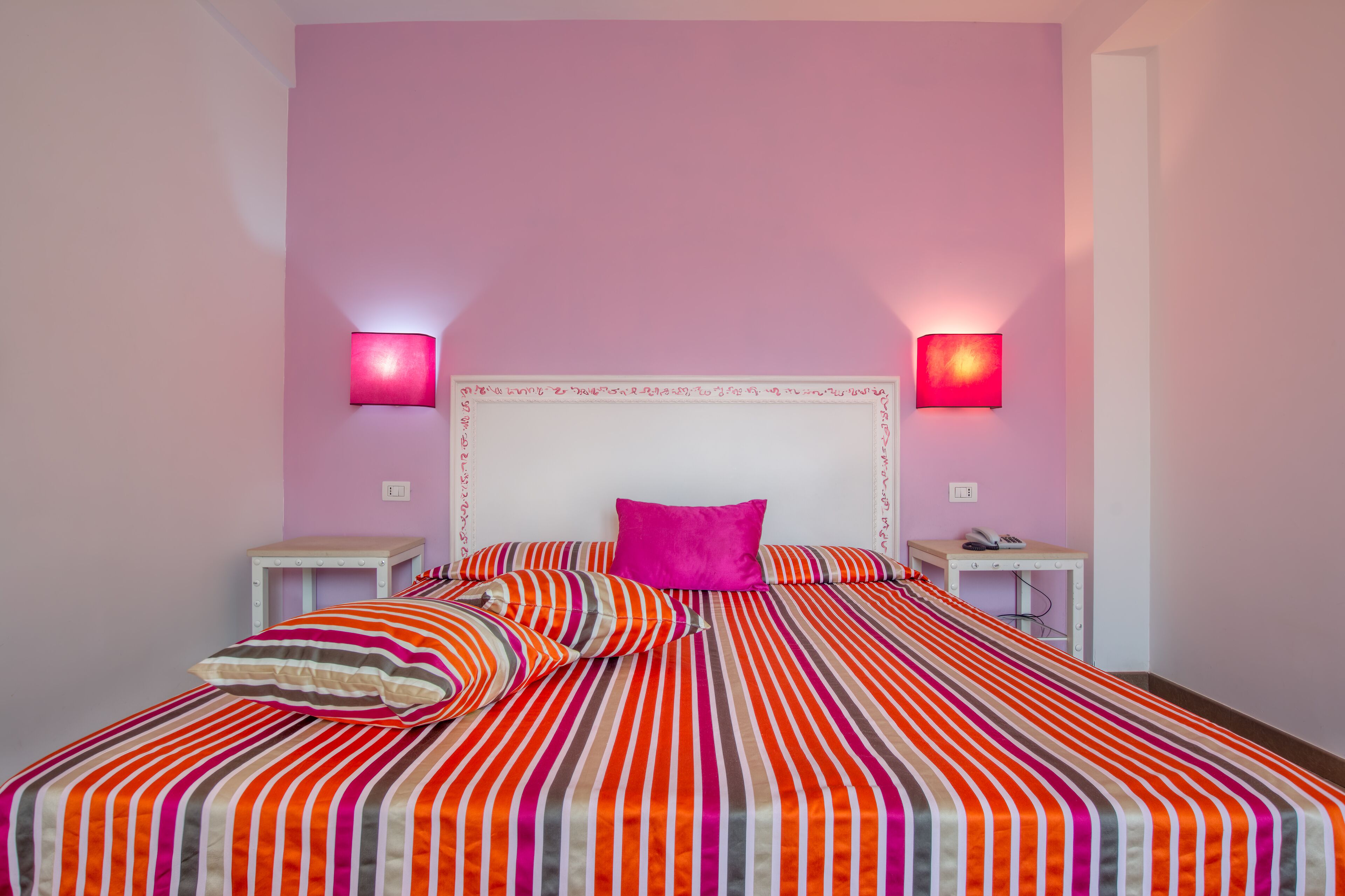 Una cama con ropa de cama a rayas y un cabecero blanco, dos mesitas de noche con lámparas, y una pared rosa.