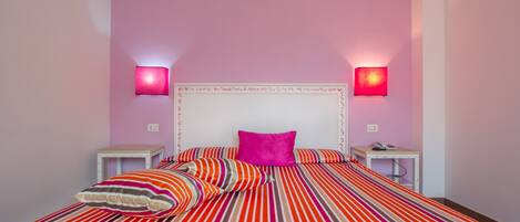 Una cama con ropa de cama a rayas y un cabecero blanco, dos mesitas de noche con lámparas, y una pared rosa.