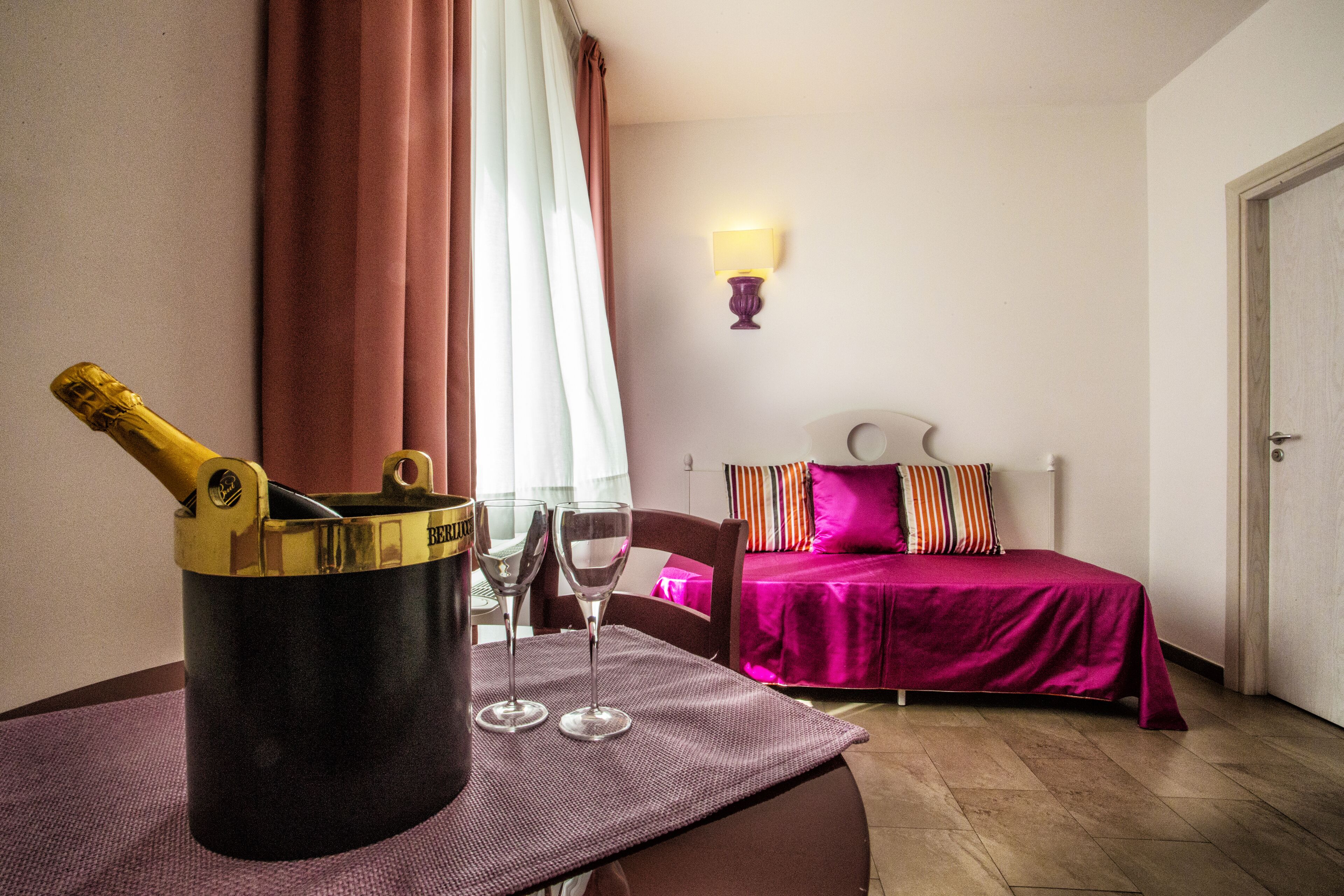 Une chambre avec un lit, une table de chevet sur laquelle se trouvent une bouteille et des verres, une lampe et une fenêtre avec des rideaux.