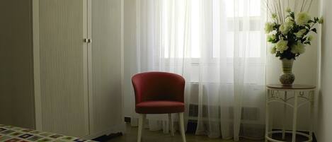 A bedroom with a bed, a red chair, a small table with a vase of flowers, and a window with sheer curtains.