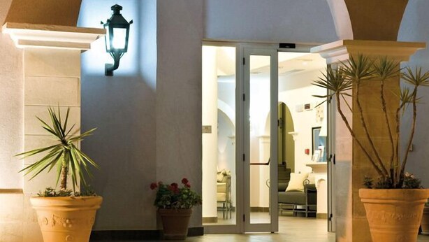 An interior courtyard with a glass door leading to a room, surrounded by arches and potted plants. - Hotel O'scià (Lampedusa)