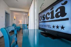 Habitación de hotel con un televisor mostrando el nombre del hotel y una valoración de tres estrellas. Hay sillas azules y una mesa en la habitación.