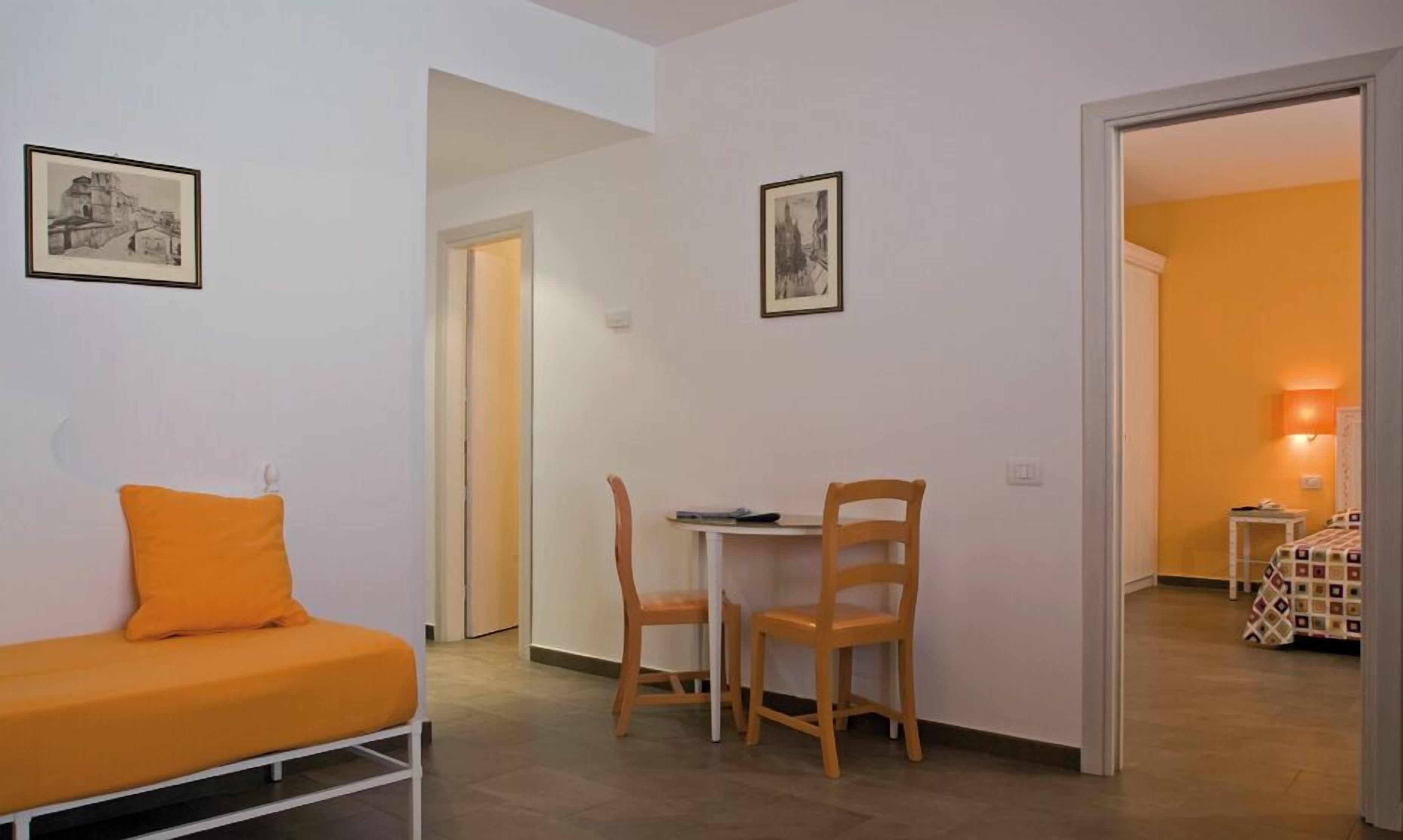 A small, bright room with a single orange sofa, a wooden chair, and a small table. There are two framed pictures on the walls and a door leading to another room.