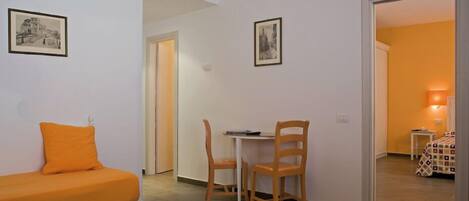 A small, bright room with a single orange sofa, a wooden chair, and a small table. There are two framed pictures on the walls and a door leading to another room.