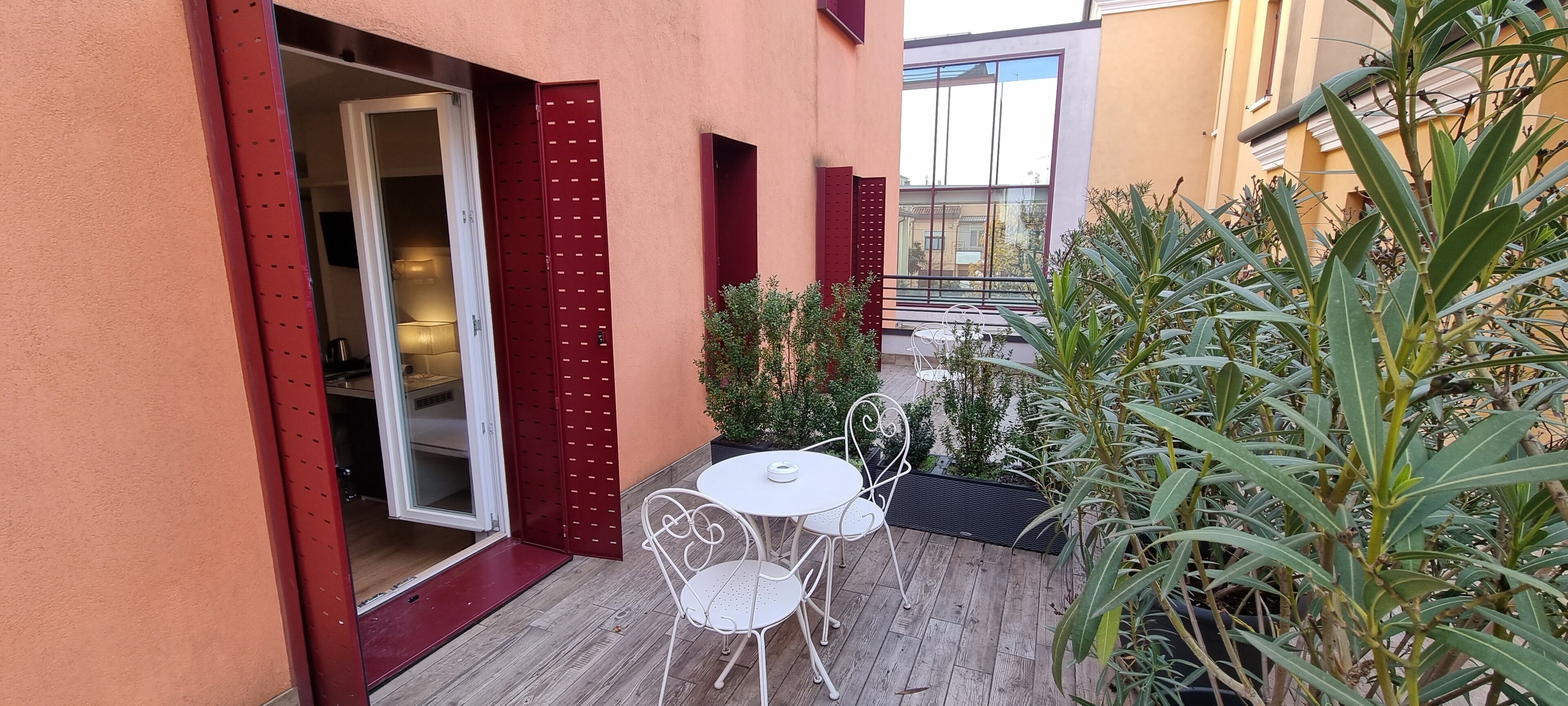 Une petite terrasse extérieure avec une table et des chaises blanches, une porte rouge et des plantes en pot.
