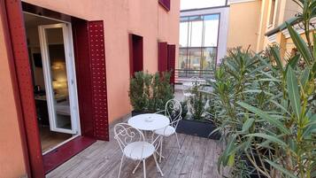 Un pequeño patio al aire libre con una mesa y sillas blancas, una puerta roja y plantas en macetas.