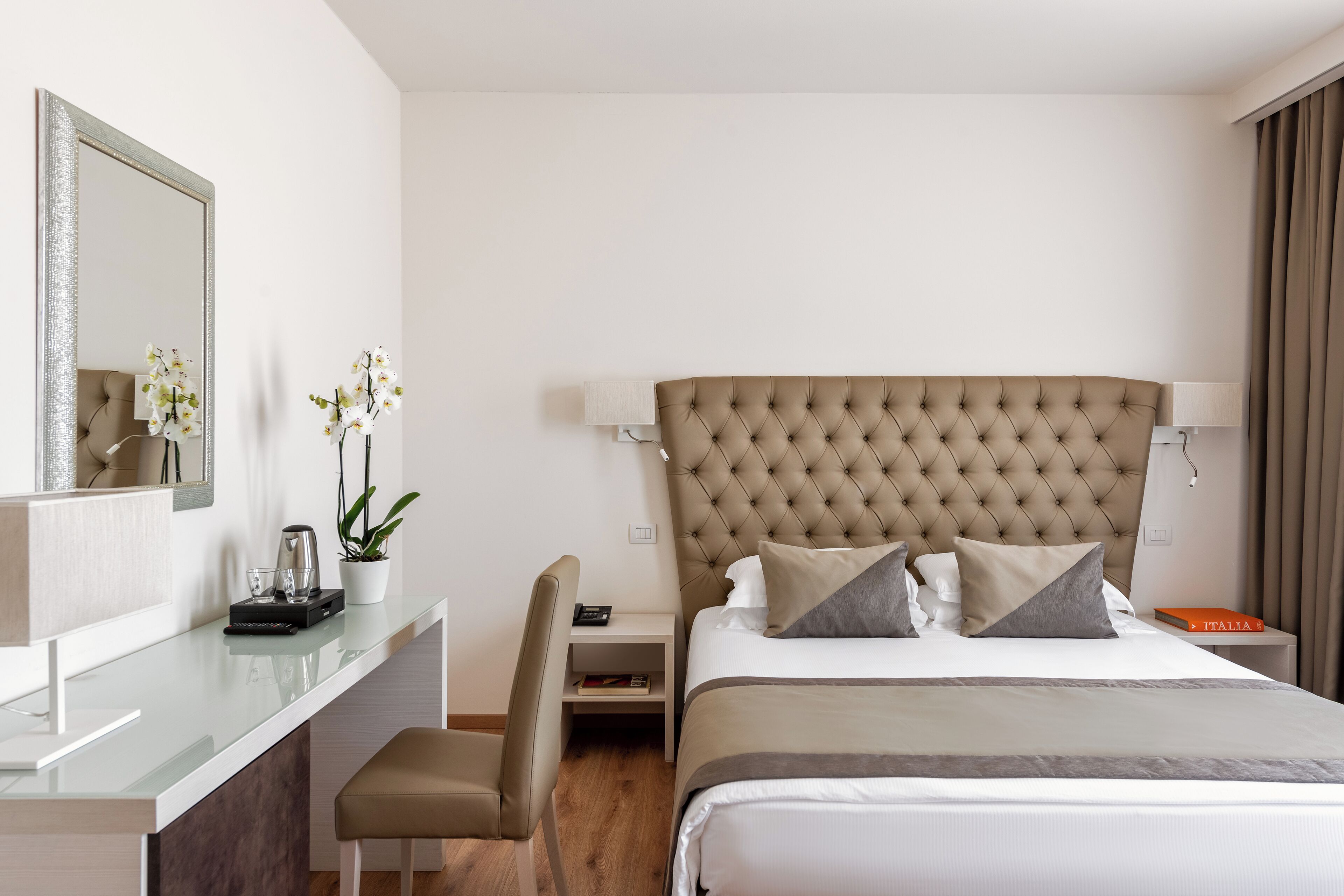 A modern hotel room with a large bed, a desk with a chair, a small table, and a mirror.