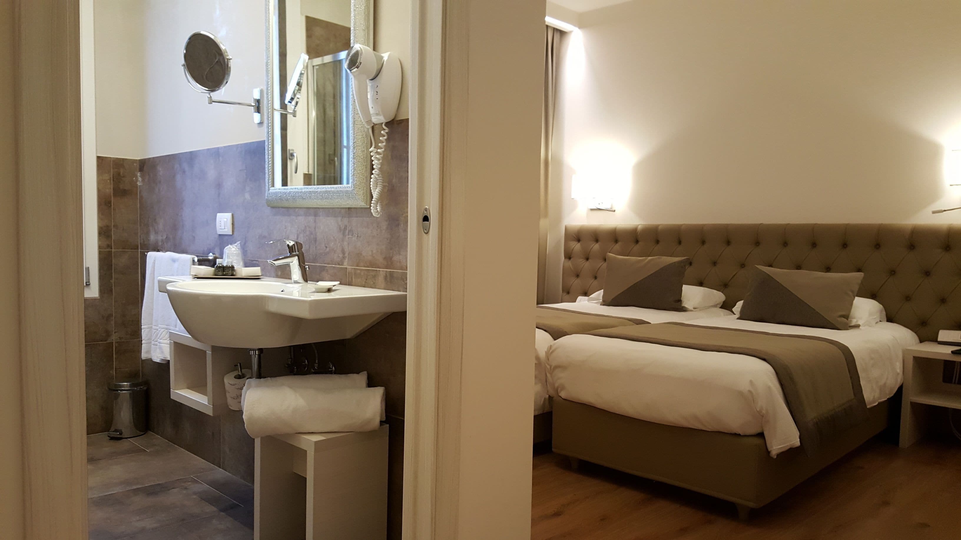 A modern bathroom with a vanity, sink, and mirror. The room features a large bed with a tufted headboard and multiple pillows.