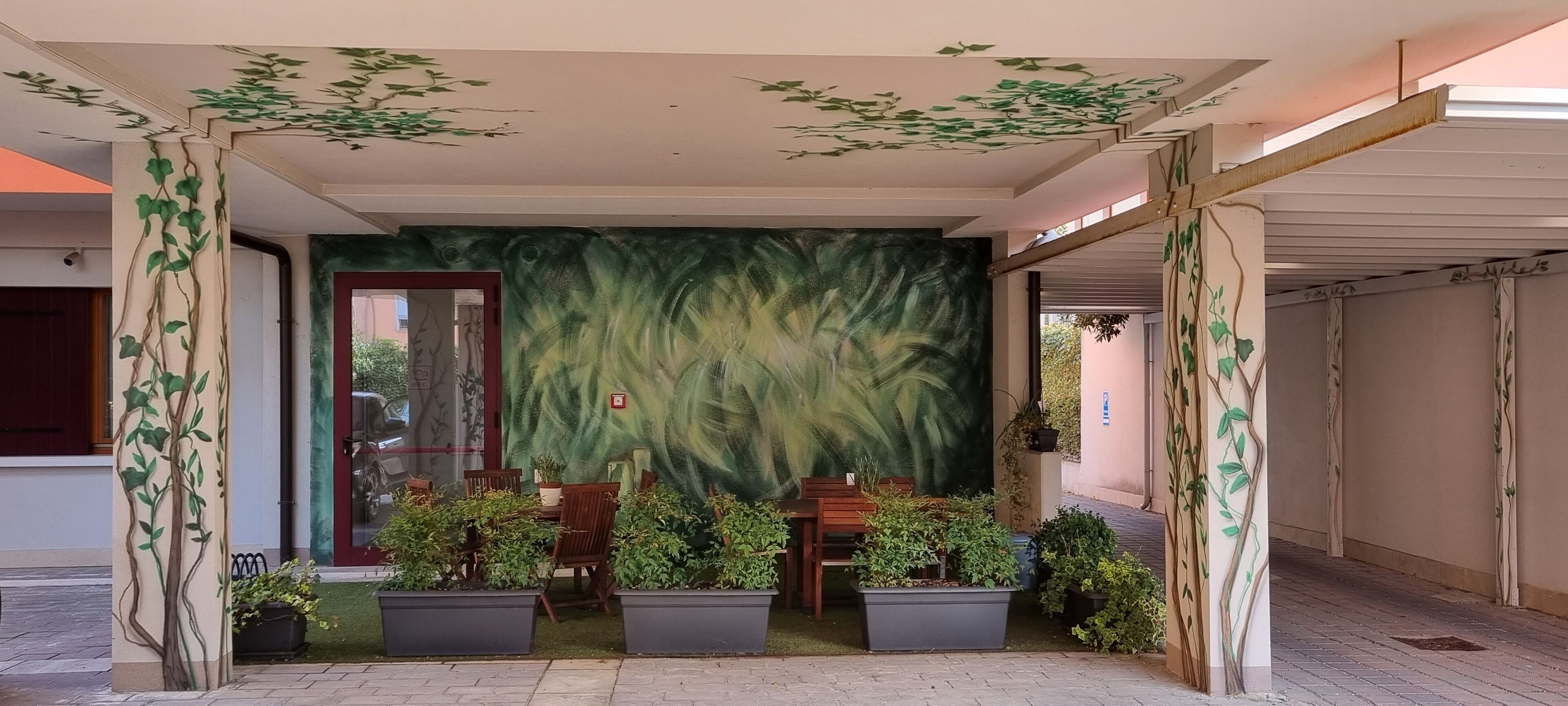 Un espacio al aire libre cubierto con mesas y sillas, rodeado de plantas en macetas y decoración mural.