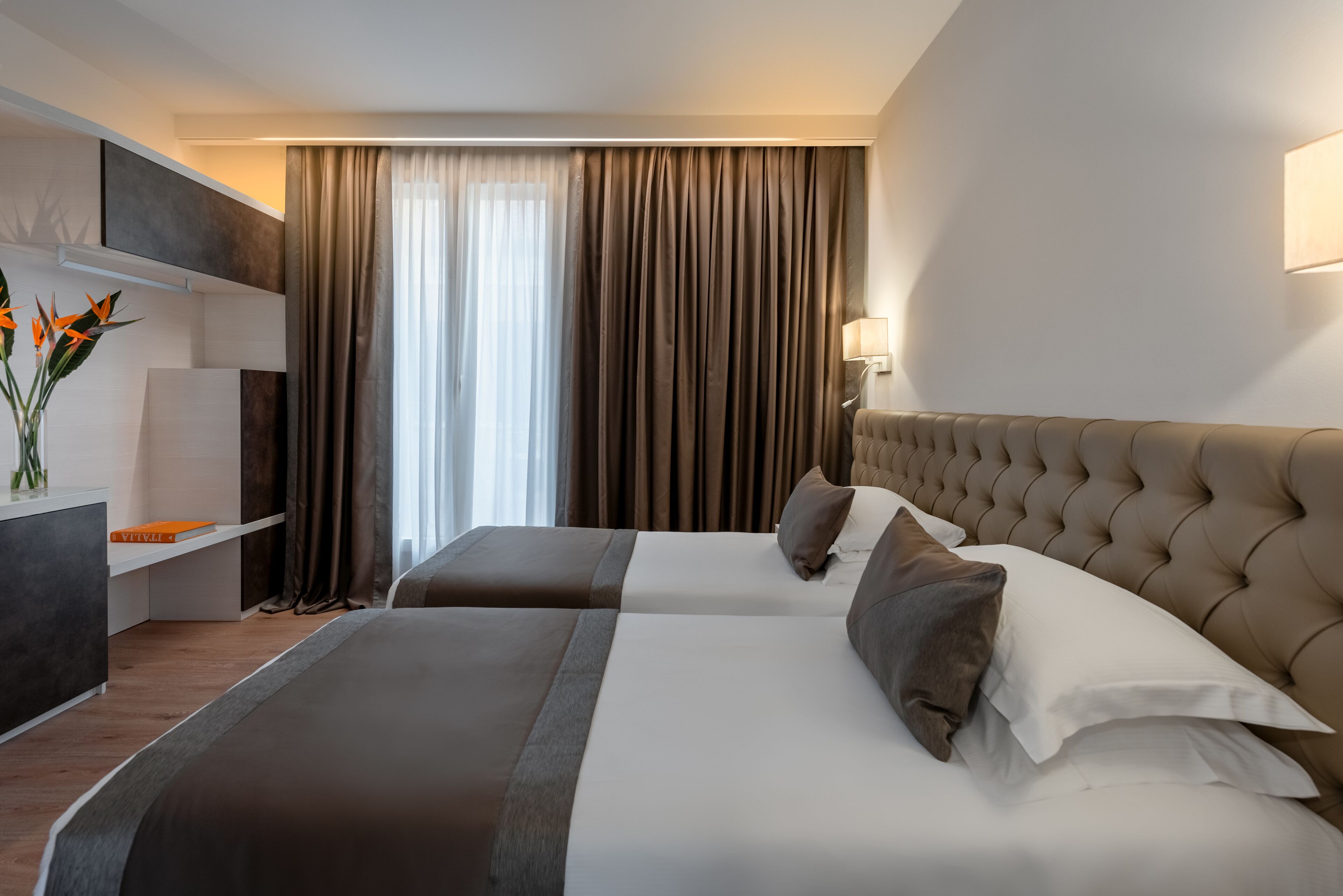 A hotel room with two beds, a bedside table, a lamp, and a large window with curtains.