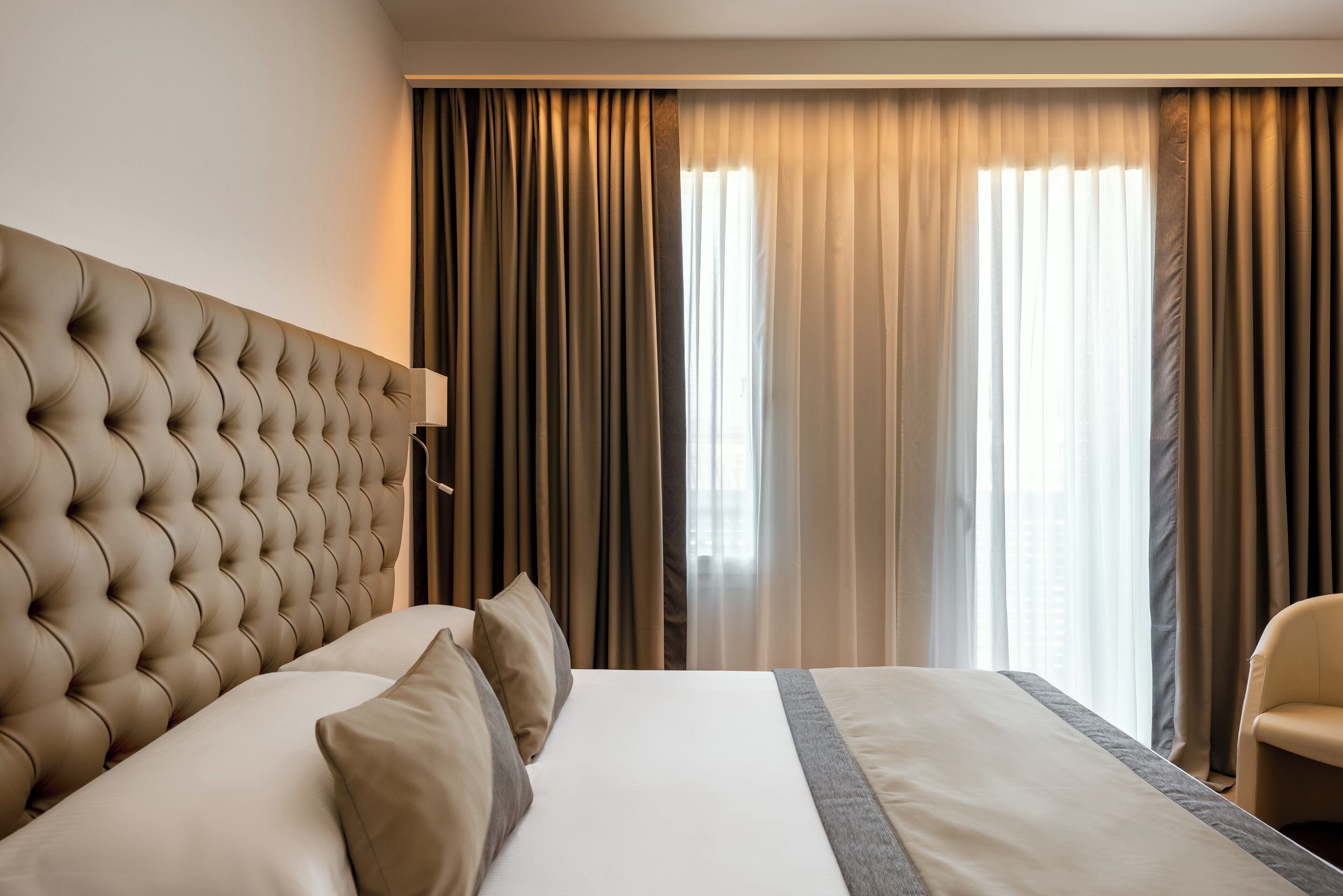 Une chambre d’hôtel avec une tête de lit capitonnée, un grand lit avec une literie beige et une vue sur la ville, visible à travers de fines rideaux.