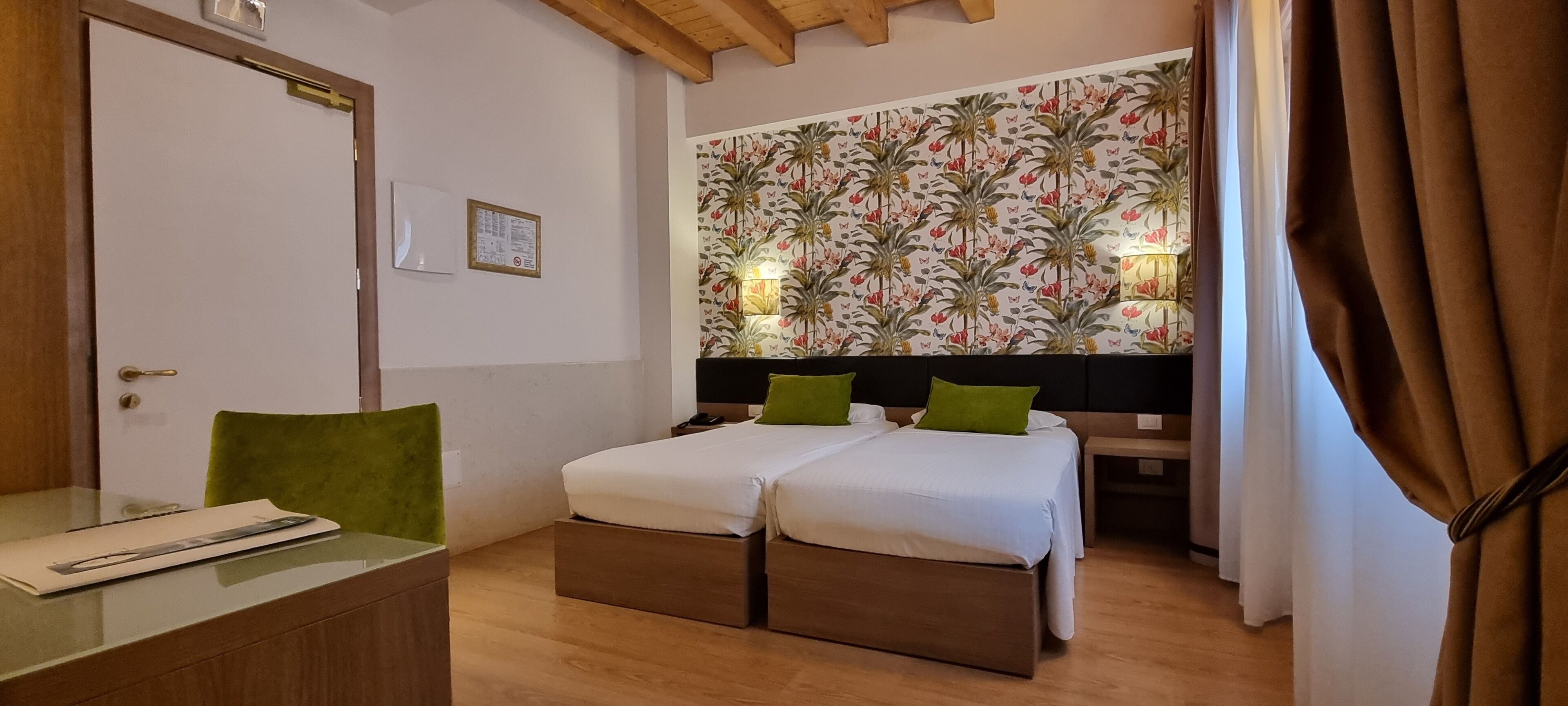 A hotel room with a wooden desk, two beds with green pillows, a floral wallpaper, and a wooden ceiling.