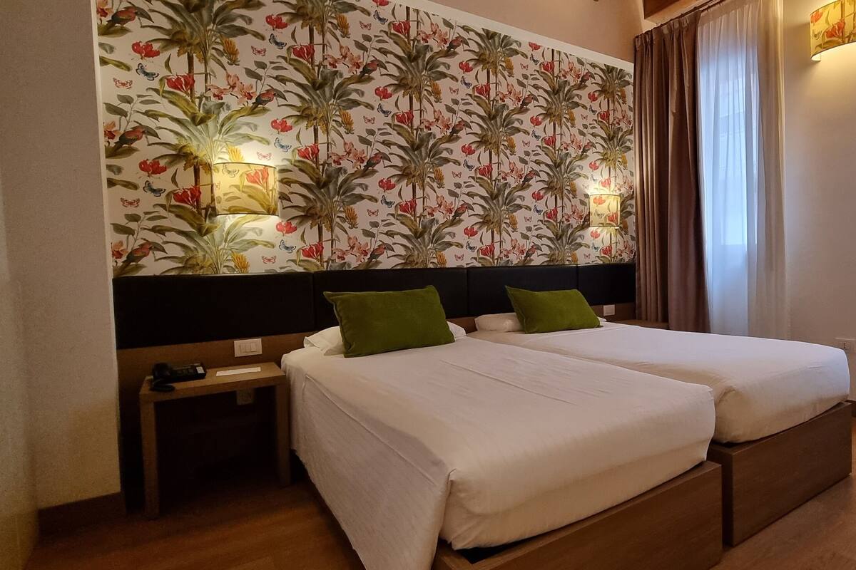 A hotel room with two beds, a floral wallpaper, a nightstand, and a window with curtains.