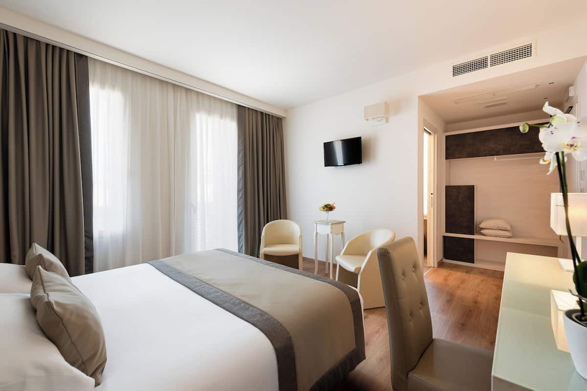 A hotel room with a large bed, two chairs, a TV, and a small table with a flower.