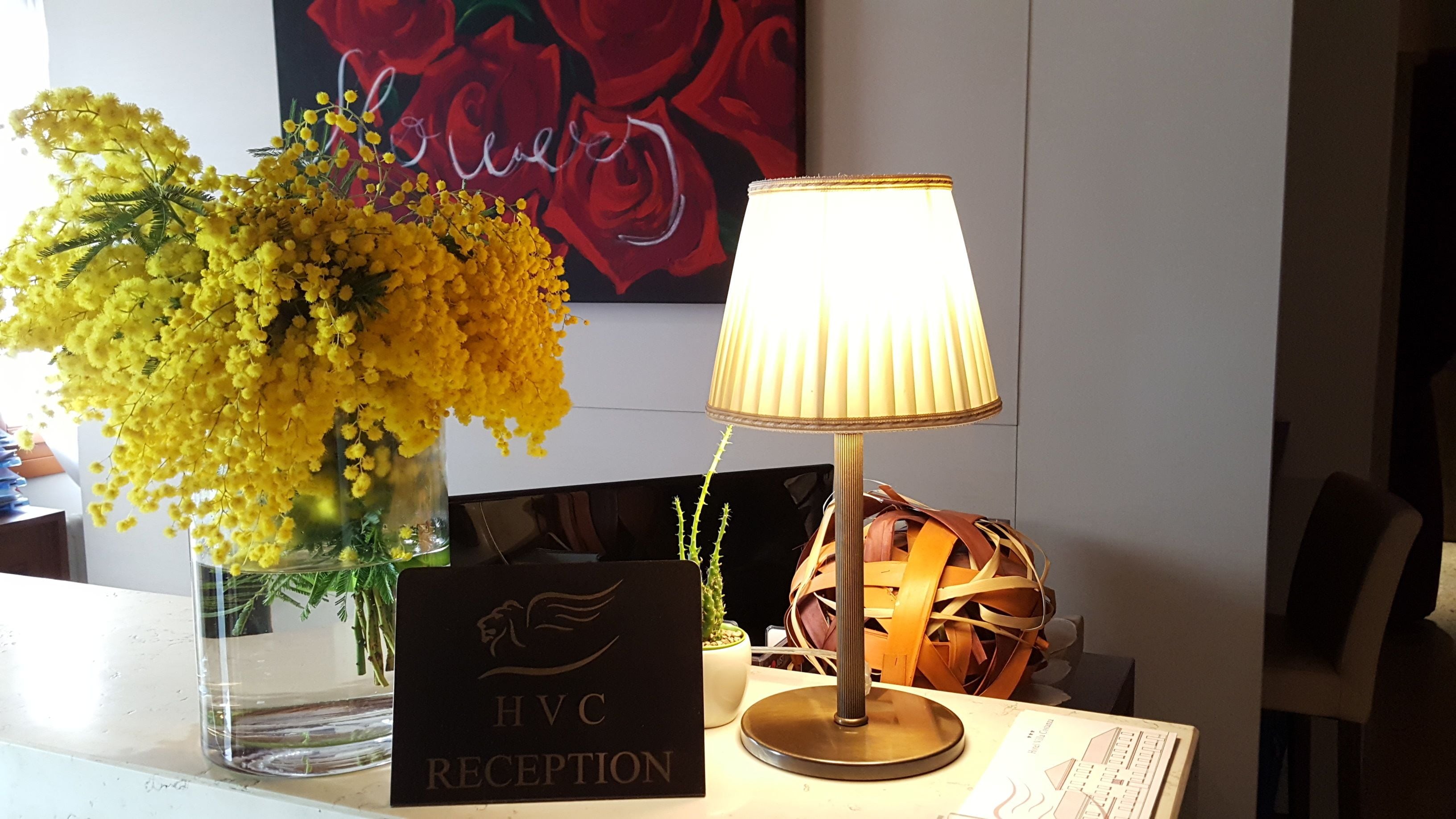 Un comptoir d’accueil orné d’un vase transparent rempli de fleurs jaunes, d’une lampe et d’une pancarte indiquant « RÉCEPTION HVC ».