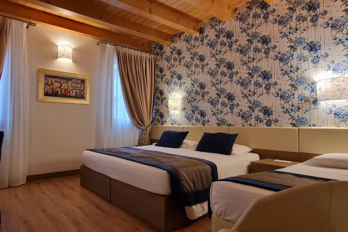 A hotel room with a wooden ceiling, a large bed with a headboard, two bedside tables, a desk with a lamp, and a wall with floral wallpaper.