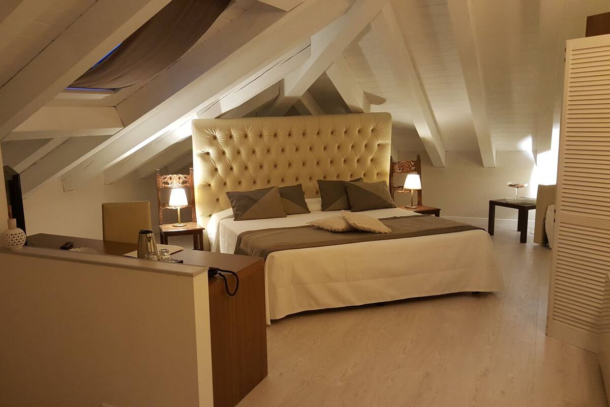A bedroom with a large bed, a desk, and a chair. There is a window with a view of the sky.