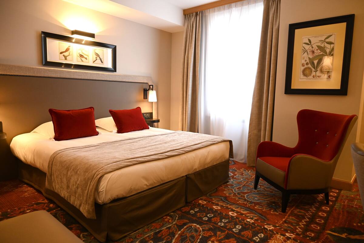 Une chambre d’hôtel avec un lit, deux oreillers rouges, une chaise rouge, une table de chevet avec une lampe et des œuvres d’art encadrées accrochées aux murs.