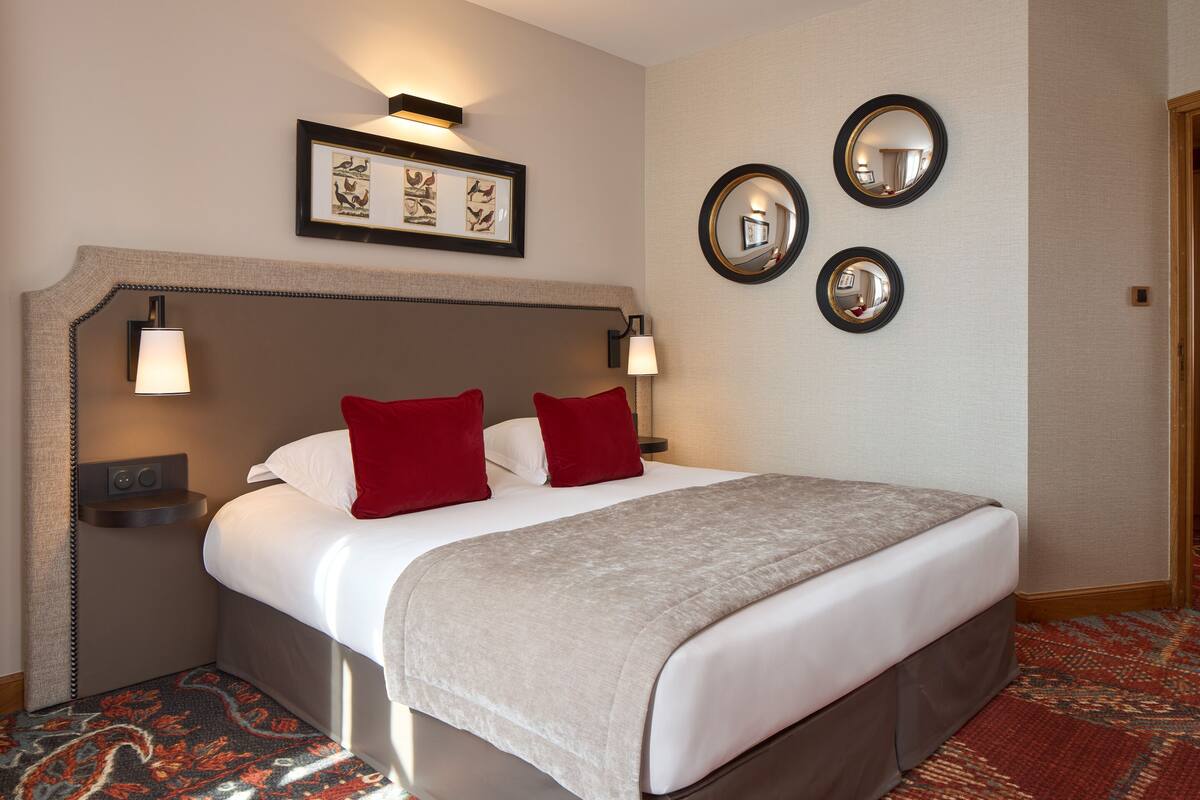 Une chambre d’hôtel avec un grand lit, deux oreillers rouges, une couverture beige, une tête de lit agrémentée de lampes murales et un tableau encadré au-dessus du lit.