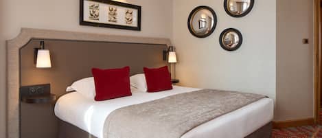 A hotel room with a large bed, two red pillows, a beige blanket, a headboard with wall-mounted lamps, and a framed picture above the bed.