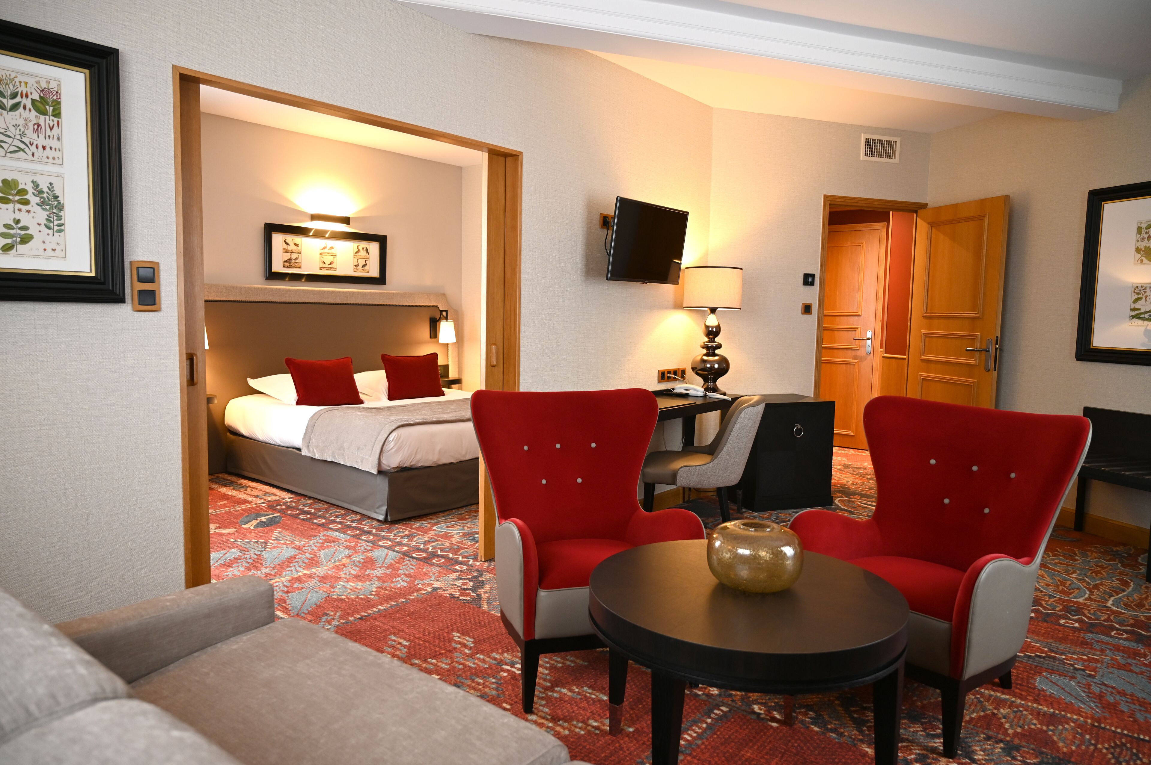 A hotel room with a bed, two red armchairs, a small table, a TV, and a patterned carpet.