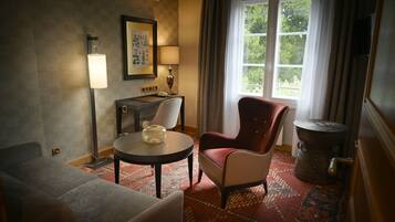 Habitación de hotel con sofá, una silla, una mesita con lámpara, un espejo decorativo y un ventanal con cortinas.