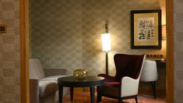 A hotel room with a patterned wallpaper, a round table with a lamp, two chairs, and a framed botanical print on the wall.